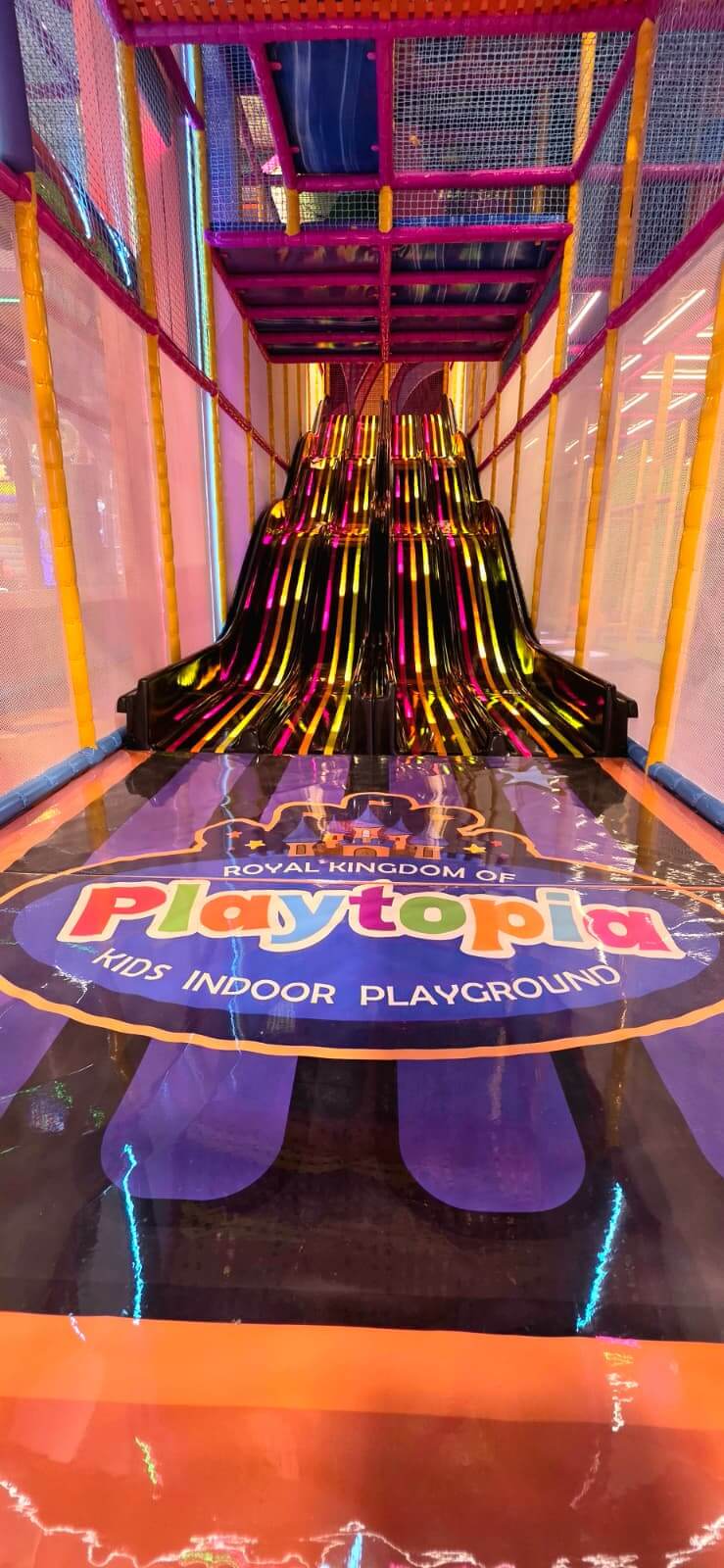 Bottom view looking up the vibrant neon-lit wavy multi-lane slide featuring the Royal Kingdom of Playtopia Kids Indoor Playground Burlington floor logo.