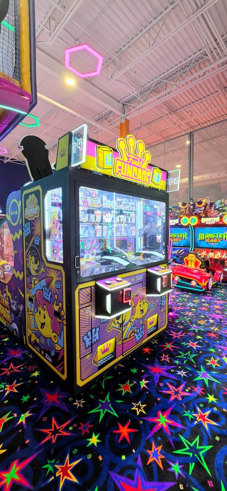 A brightly lit Fun Zone claw machine crane game full of plush toys next to racing simulator games at Playtopia Burlington.