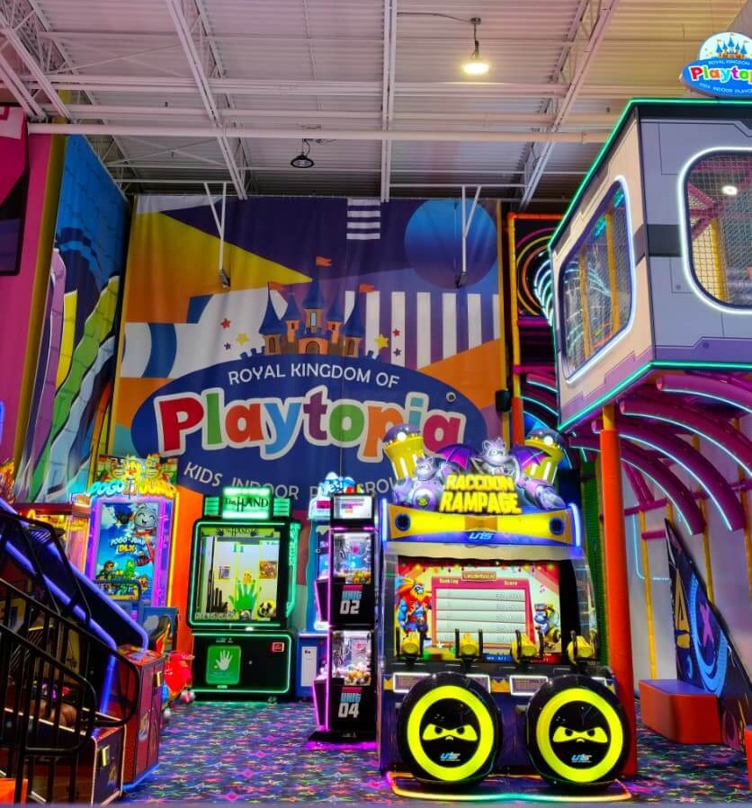 Vibrant arcade gaming zone under a high ceiling showing cabinet ticket games and a massive Royal Kingdom of Playtopia wall logo in Burlington.